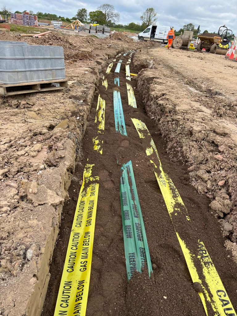 Close-up view of an excavated utility trench featuring yellow gas and electric caution tapes alongside green telecommunications warning tape