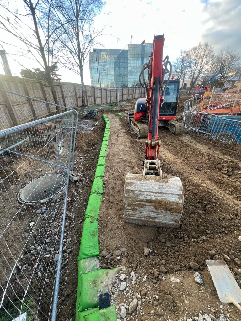 Red excavator performing commercial groundworks and drainage installation alongside a green retaining wall in Preston Deanery