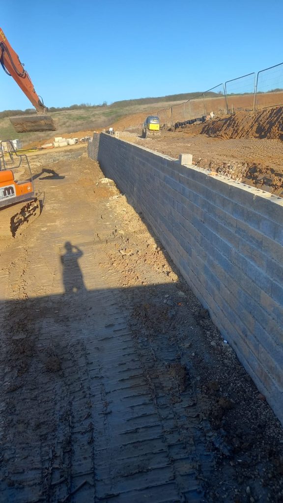 Heavy machinery excavating ground alongside newly built Allan Block retaining wall in Wellingborough
